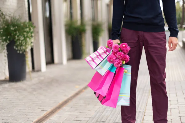 Elegante hombre sosteniendo flores y rosa, bolsas de compras azules cerca del restaurante. Día de la mujer. Día de San Valentín. Listo para fiesta de cumpleaños o cita romántica. Copiar espacio. Manojo de bolsas de regalo de papel —  Fotos de Stock