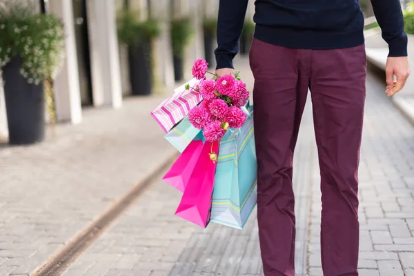 Hombre romántico con flores rosadas y bolsas de compras sobre fondo de ladrillo. Día de la mujer. Día de San Valentín. Listo para fiesta de cumpleaños o cita romántica. Copiar espacio. Manojo de bolsas de regalo de papel —  Fotos de Stock
