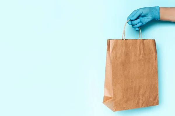 Hand in medical glove gives craft box over blue background. Contactless delivery, online shopping concept. Banner, copy space. Safe package during Coronavirus pandemic. Zero waste lifestyle.