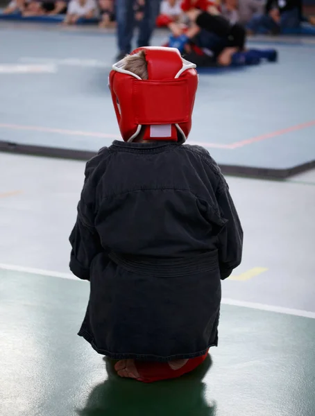A young athlete boy in boxing gloves and a protective headgear looks at the tatami with the fighters.