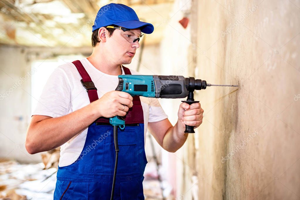 Construction worker in protection glasses and uniform with perforator drilling the wall indoors. Man with drill