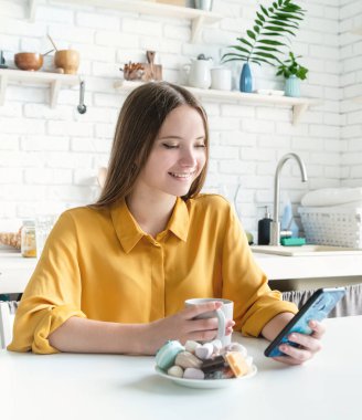 Mutfakta yemek yerken akıllı telefonunu kullanan gülümseyen genç bir kadın.