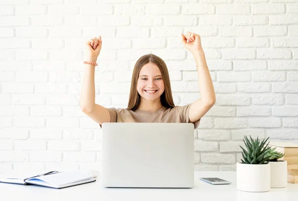 Excited teen girl study using her laptop with her arms raised. Happy teenager accomplished her homework, got the high mark on exam, excited student