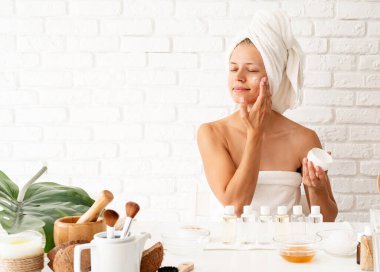 Spa and beauty concept. Happy young woman wearing white bathrobes towels on head doing spa procedures applying creme on her face in spa beauty salon clipart