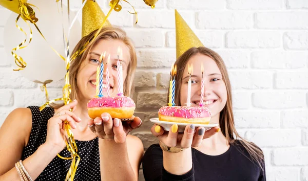 Dos mujeres jóvenes felices en sombreros de cumpleaños celebrando globos de cumpleaños, la celebración de donas con velas sobre fondo de pared de ladrillo blanco —  Fotos de Stock