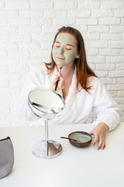 Spa ve sağlık. Doğal kozmetikler. Kişisel bakım. Bornoz giyen, aynaya bakan, kilden maske takan beyaz bir kadın.