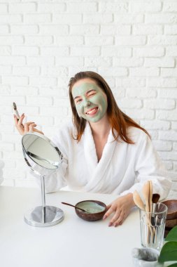 Spa ve sağlık. Doğal kozmetikler. Kişisel bakım. Genç, beyaz, bornoz giymiş, kilden maske takan, komik suratlı bir kadın.