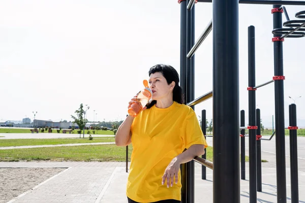 Mujer mayor sonriente bebiendo agua después del entrenamiento al aire libre en el campo de deportes — Foto de Stock