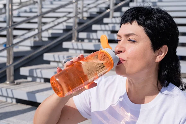 Sport and fitness. Senior sport. Active seniors. Smiling senior woman drinking water after workout outdoors on urban background