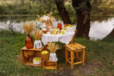 Açık havada, sakin bir su kütlesinin hazırladığı süslü bir masa. Masa beyaz bir bezle kaplı, güzel bir hasat gösterisiyle dolu küçük beyaz balkabakları, taze sarı elmalar,