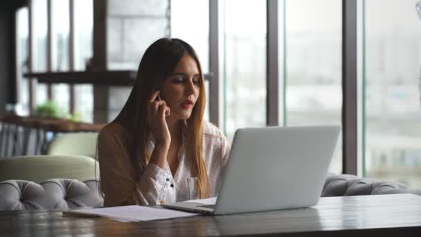 Frumoasă femeie tânără care lucrează cu laptop și vorbește pe smartphone vorbind cu clientul care stă lângă fereastră — Videoclip de stoc