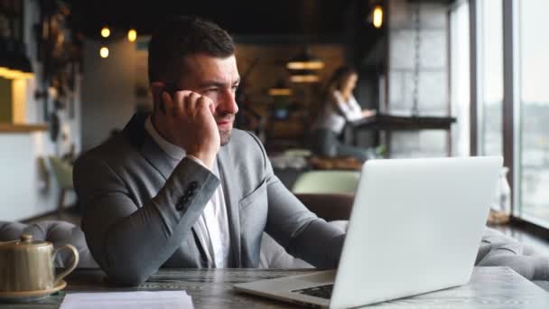 Jonge zakenman die op laptop werkt en praten over telefoon, aan tafel aan op café — Stockvideo