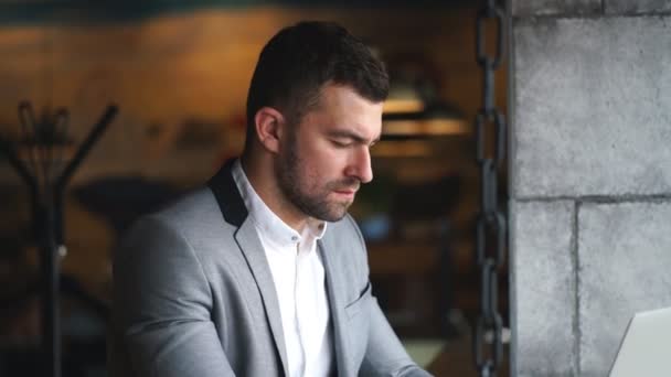 Un joven hombre de negocios con un traje gris y una camisa blanca trabaja en un ordenador portátil en un café — Vídeos de Stock