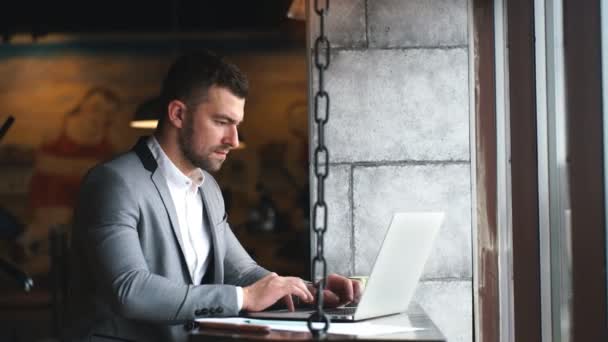 Вид збоку серйозного бородатого чоловіка-підприємця, що сидить з ноутбуком в кафе, робить віддалену роботу — стокове відео