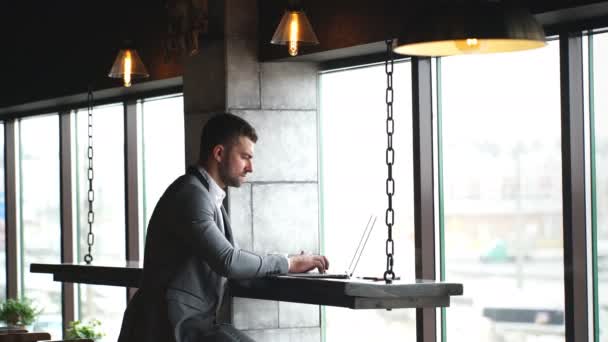 Ein junger gutaussehender Geschäftsmann checkt Dokumente und tippt auf einem Laptop, der in einem Café am Fenster steht — Stockvideo