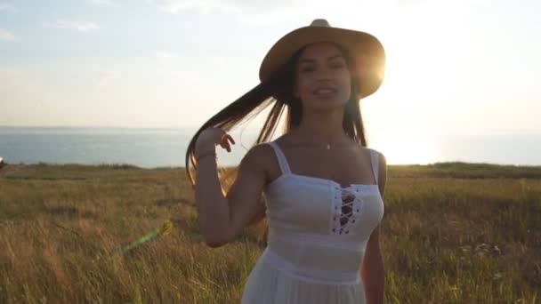 Encantadora morena en un vestido de luz blanca y sombrero girando en el campo — Vídeos de Stock