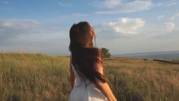 An einem sonnigen Tag läuft eine Frau mit einem weißen Kleid vor einem Naturhintergrund an einem Weizenfeld entlang — Stockvideo