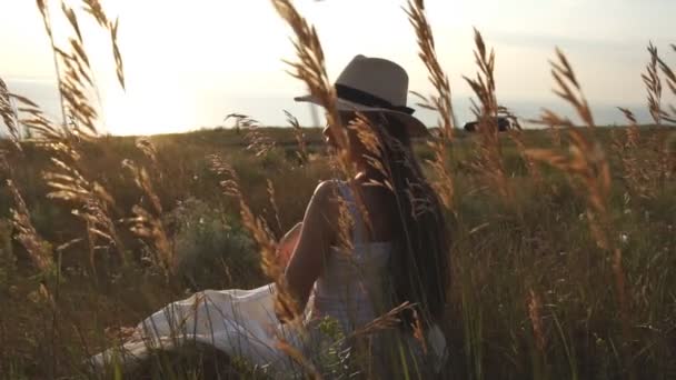 Brunette in een stro hoed zit in het veld en de zonsondergang bewondert — Stockvideo