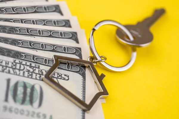 The concept of buying a private house or apartment. Mortgage registration. The accumulation of money for real estate. Metal key chain in the shape of a house with a metal key lying on the pack of hundred US dollars on a yellow background. Close up.