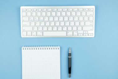 Defter beyaz kağıt ve kalem, mavi zemin üzerine beyaz kablosuz klavye ile kontrol.