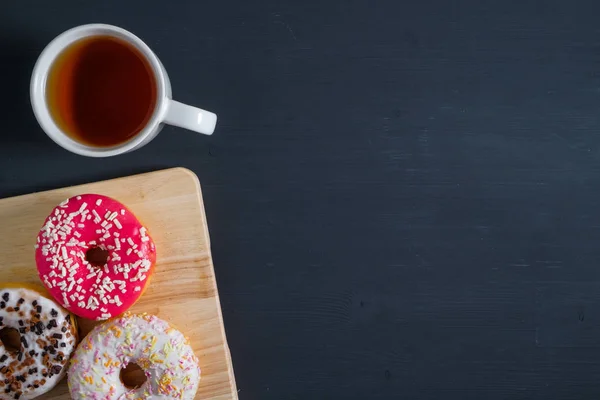 Beyaz, pembe ve kahverengi donutlar, sol tarafında bir fincan çay ve siyah ahşap arka plan.. — Stok fotoğraf