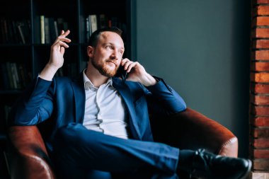 takım elbiseli başarılı işadamı modaya uygun bir ofisin sandalyesinde oturur ve akıllı telefonda bir iş planını tartışır.