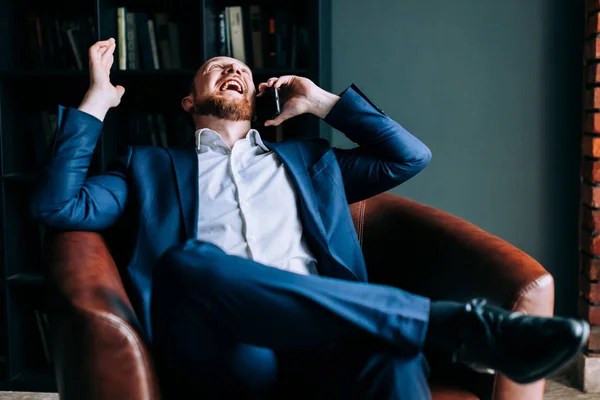 Successful businessman in a suit sits in a chair of a trendy office and laughs out loud.