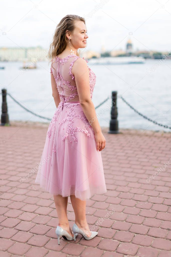 Young curly Caucasian woman in rose pink dress turned her back on embankment.