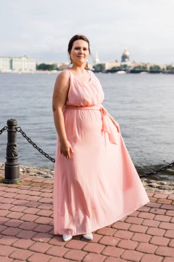 Uzun pastel pembe elbiseli esmer kafkas modeli rıhtımda kameraya bakıyor.