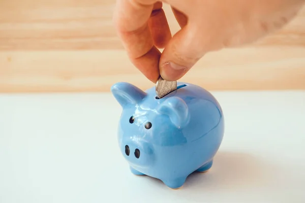 Hand is putting coin in blue piggy bank on wooden and white background. — Stock Photo, Image