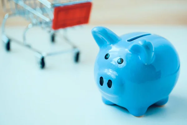Close-up van blauwe Piggy Bank op achtergrond klein speelgoed winkelmandje. — Stockfoto