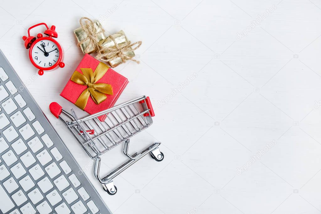 Christmas online shopping concept. White keyboard with red clock, trolley and gift on a wooden background. Flat lay.