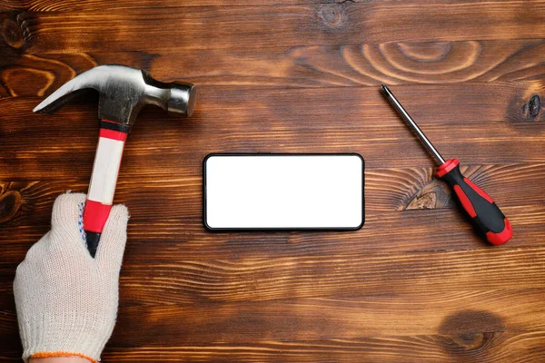 Hand of a repairman in a glove with a hammer and a smartphone next to it with copy space, screwdriver on a wooden background. Top view