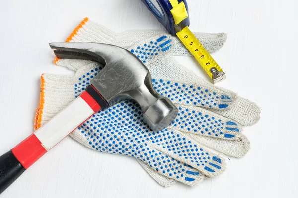 Hammer, measuring tape on work gloves and a white wooden background.