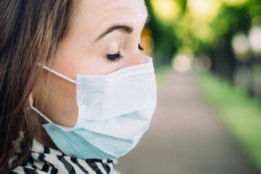 Park arka planında gözleri kapalı bir şekilde tıbbi maske takan 30 yaşında beyaz bir kızın portresi..