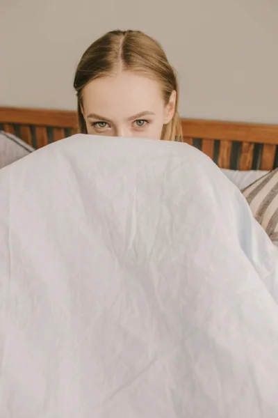 Retrato de uma menina bonita está descansando na cama em seu quarto e tela-se atrás do cobertor azul-claro — Fotografia de Stock