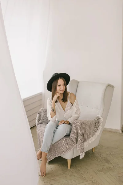 Young woman in hat at home sitting on modern chair in front of window relaxing in her light living room — Stock Photo, Image