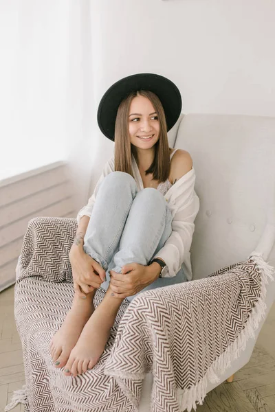 Young woman in hat at home sitting on modern chair in front of window relaxing in her light living room — Stock Photo, Image