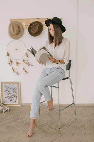 Young brunnete girl in black hat, white shirt and blue jeans is reading a magazine in the light boho room — Stock Photo, Image