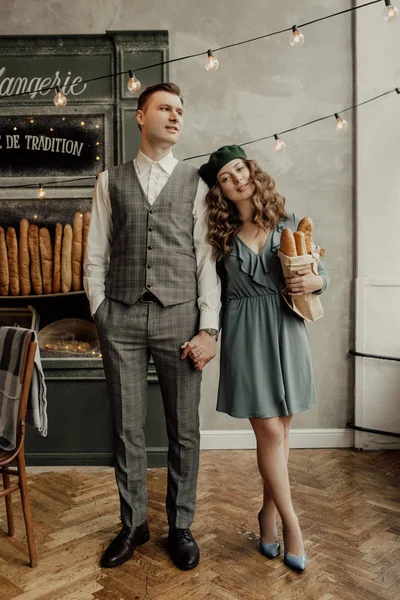 Genç Fransız Cafe arka plan üzerinde şık Romantik Fransız çift duruyor seviyorum. Şık adam kız ellerini tutar. O yeşil bere takıyor ve baget tutar. Lighs arkalarında vardır