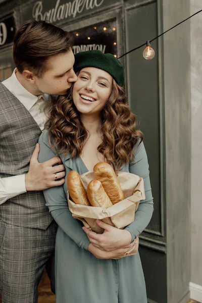 Young stylish romantic French couple stands on the french cafe background. Stylish man is kissing the girl in her cheek. She wears green beret and holds baguettes. Lighs are behind them — Stock Photo, Image