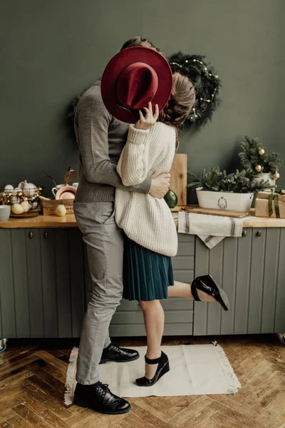 Stylish couple in love is kissing in the kitchen on Christmas eve and hiding behind the hat — Stock Photo, Image