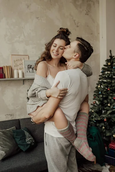 L'homme tient sa petite amie heureuse et l'embrasse dans la joue. Arbre de Noël est derrière eux et la maison ambiance chaleureuse autour — Photo