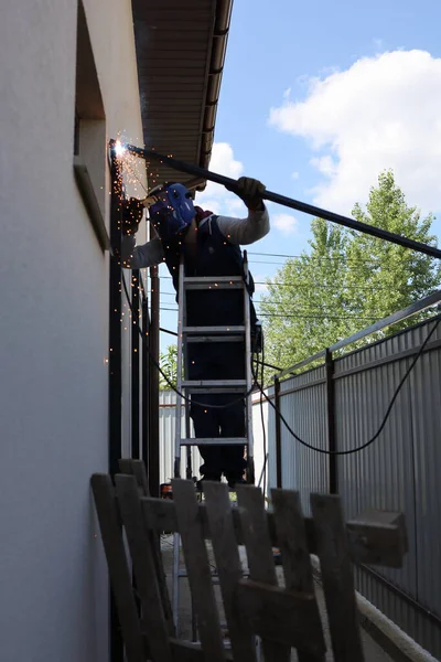 A male welder in a protective face mask and gloves welds a piece of metal to a fence. A novice welder works with a fence in a private house
