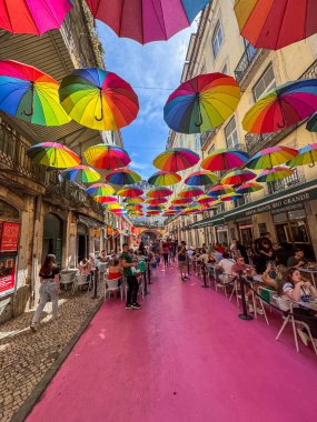 Tavandan sarkan bir sürü şemsiyesi olan renkli bir cadde. İnsanlar masada oturup yemek yiyorlar.
