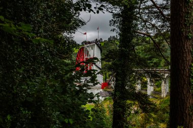 Laxey, Man Adası 08.05.2023 Büyük kırmızı yel değirmeni bir ormanın ortasında. Yel değirmeninin etrafı ağaçlarla çevrili ve üstünde Man Adası bayrağı var. -