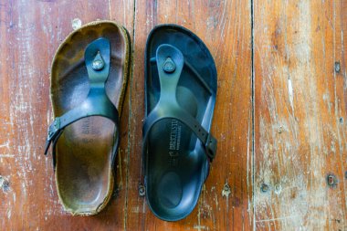 İki parmak arası terlik, biri kahverengi ve biri siyah, tahta bir yüzey üzerinde sergilenir. Kahverengi flop yıpranmış görünürken, siyah flip flop yepyeni görünüyor.