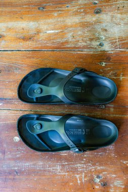 İki siyah parmak arası terlik ahşap bir masada yatıyor. Ayakkabılar siyah ve ayak bileğine takılan bir kayışı var.