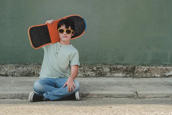 happy kid with skateboard and sunglasses sitting on the street