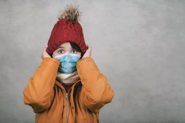 kid with medical mask wearing winter hat and scarf over gray background